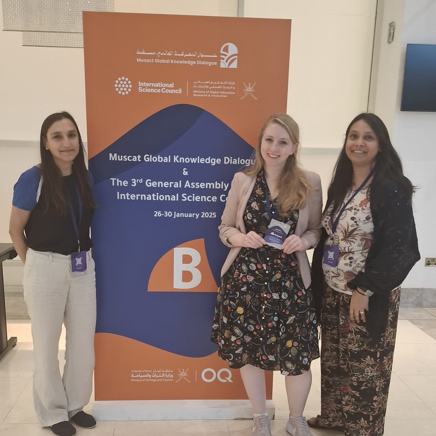 Sandeep, Amy, and Dhruti stand in front of a banner celebrating the International Science Conference.