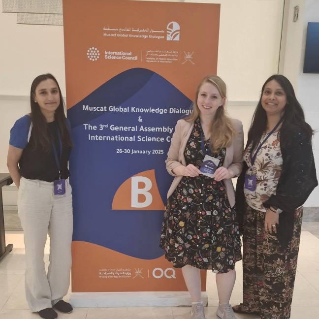 Sandeep, Amy, and Dhruti stand in front of a banner celebrating the International Science Conference.