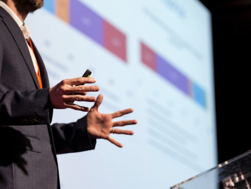 Man presents at a conference with a lectern and a screen showing graphs behind him