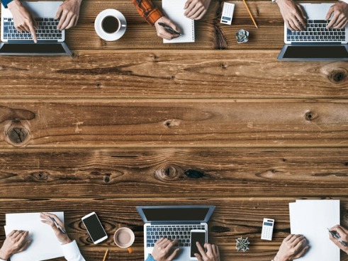 Long table with people on either side, birds-eye view where you can see laptops, pens, papers, pencils, hot drinks and phones of the different participants.