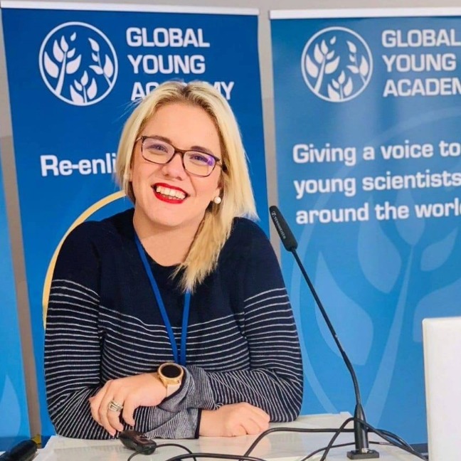 Woman smiles at camera on a panel with a microphone for the Global Young Acacdemy