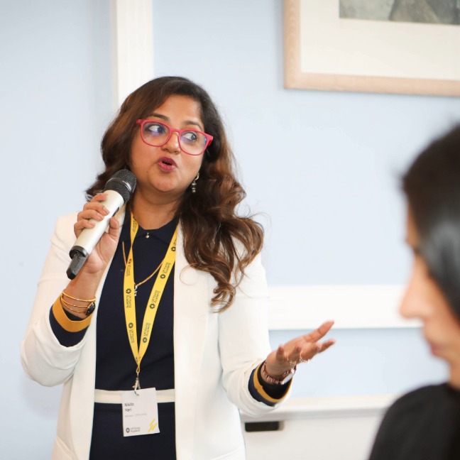 Member Nikita Hari speaks into a microphone to a room of people, and a UK Young Academy lanyard around her neck