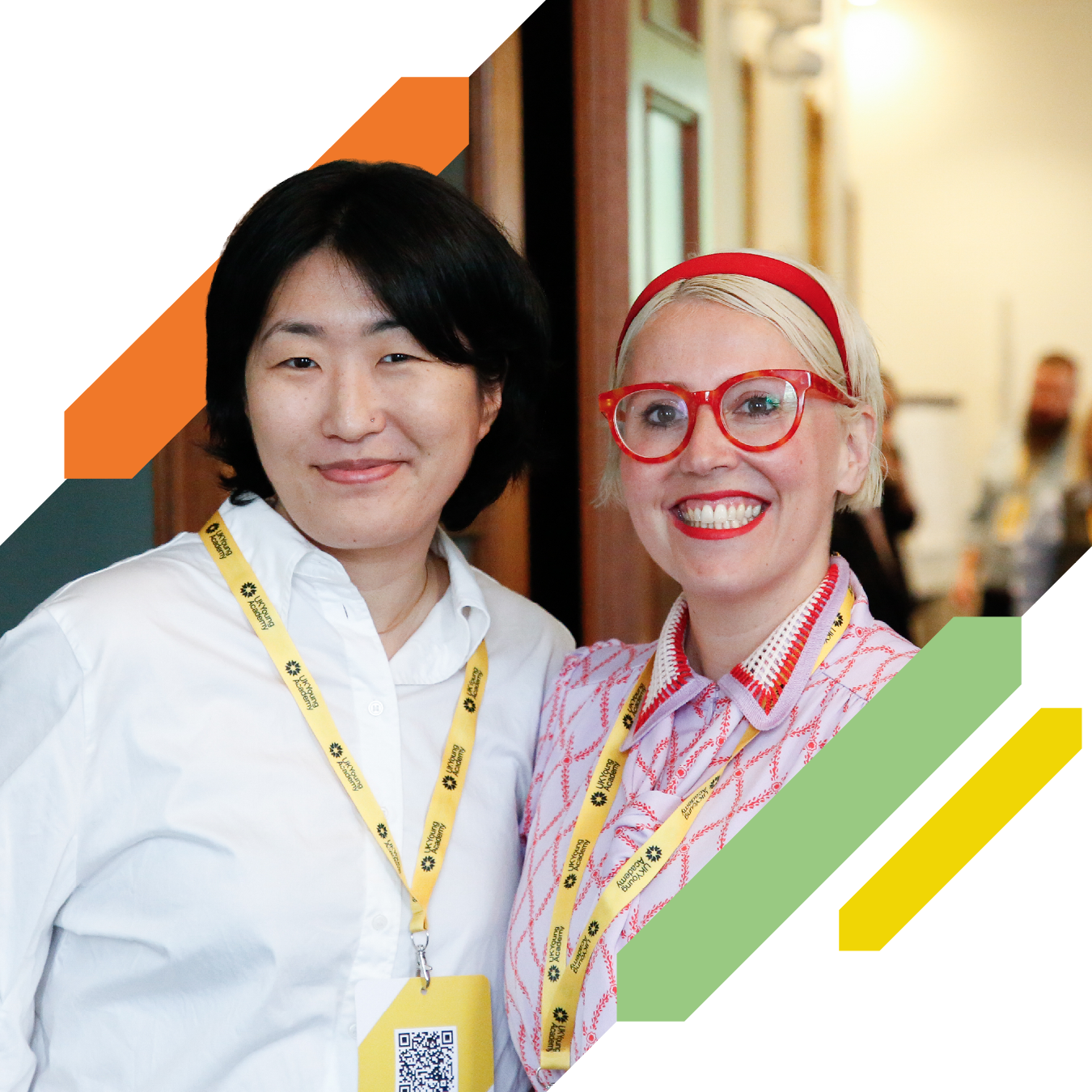 Two conference attendees wearing lanyards stand side by side facing the camera. Decorative yellow, green, orange, and grey graphic elements frame the image.