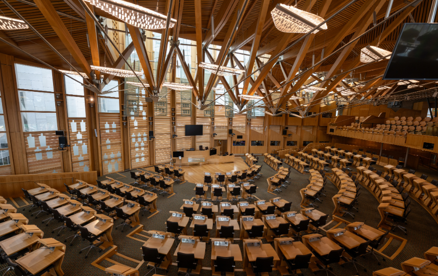 The debating chamber of the Scottish Parliament at Holyrood, with curved rows of desks and seats beneath a distinctive timber roof.