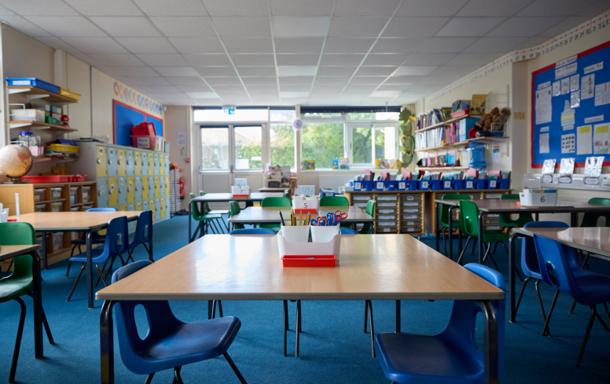 Empty UK primary school classroom with desks, chairs, storage units and learning materials arranged around the room.
