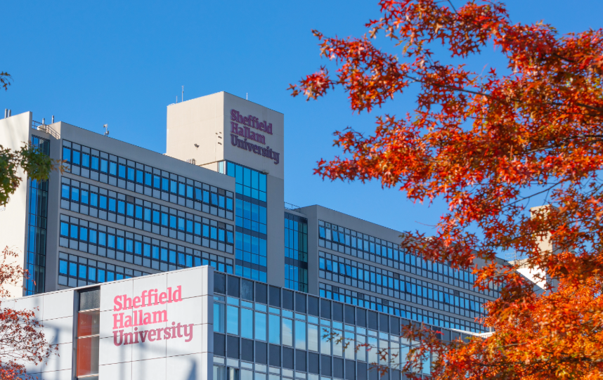 Sheffield Hallam University campus buildings with large glass windows, framed by red autumn foliage against a clear blue sky.
