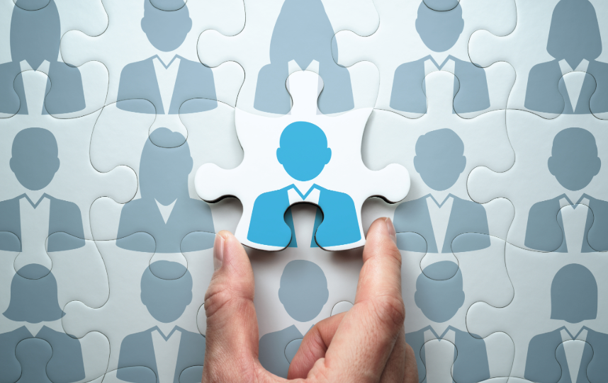 Hand placing a blue puzzle piece with a person icon into a larger puzzle made up of similar people-shaped pieces.