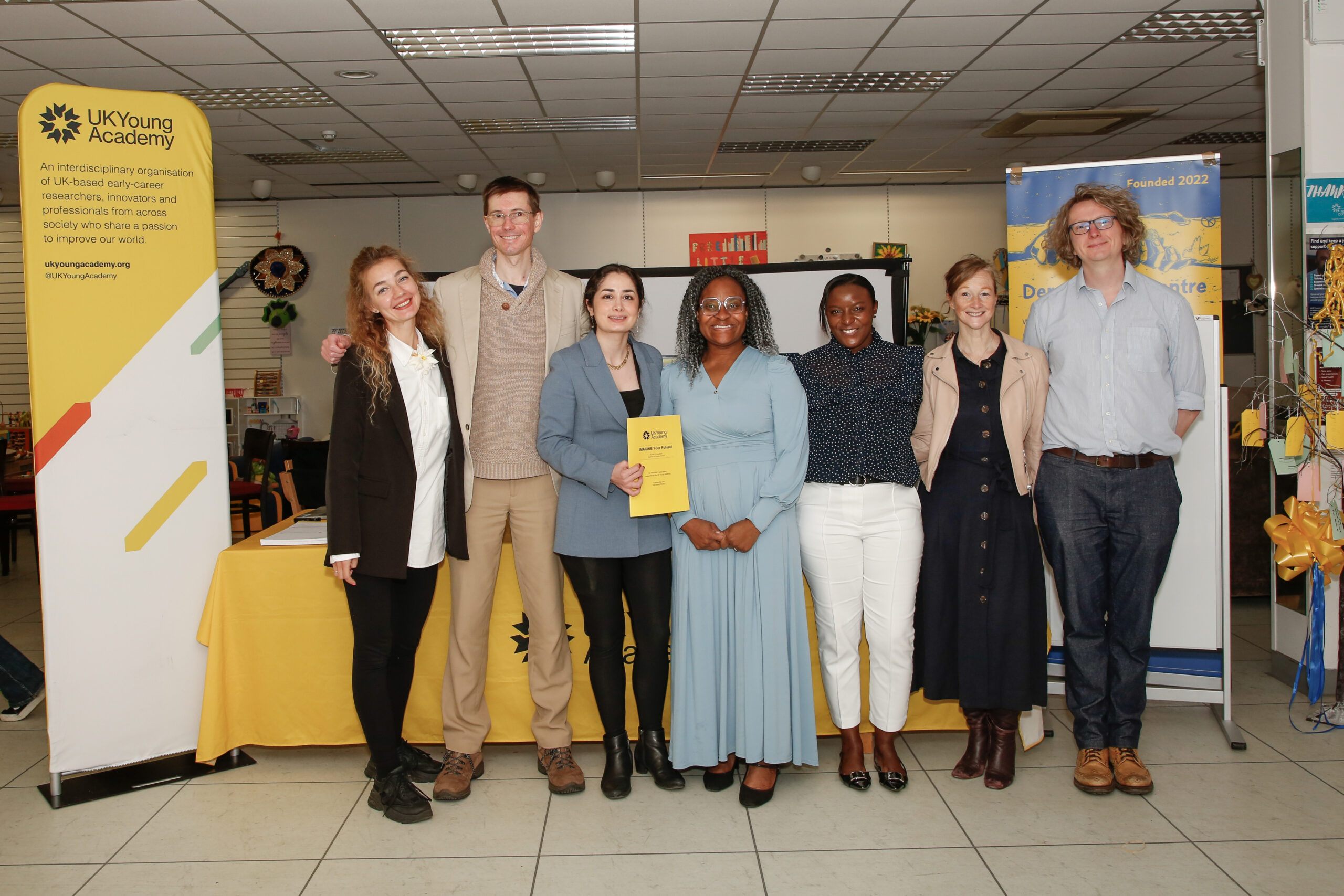 Seven members of the IMAGINE project team pose beside UK Young Academy display banners and an information table during an outreach event.