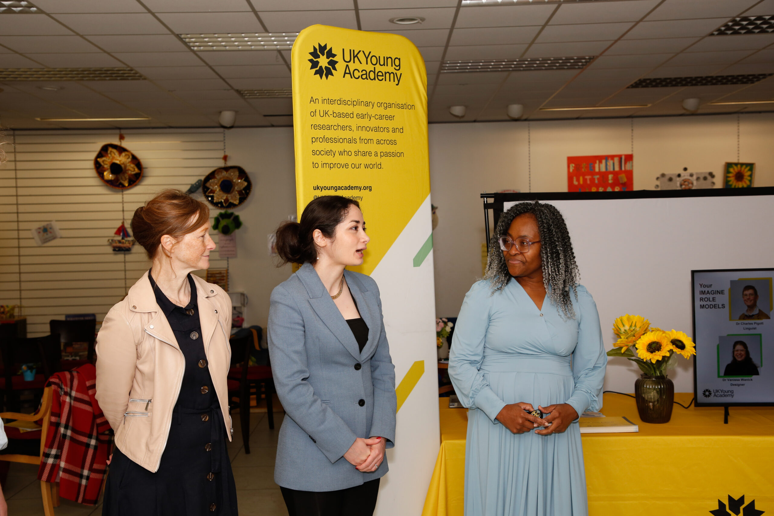 Three speakers stand beside a UK Young Academy banner and display table during an outreach event.
