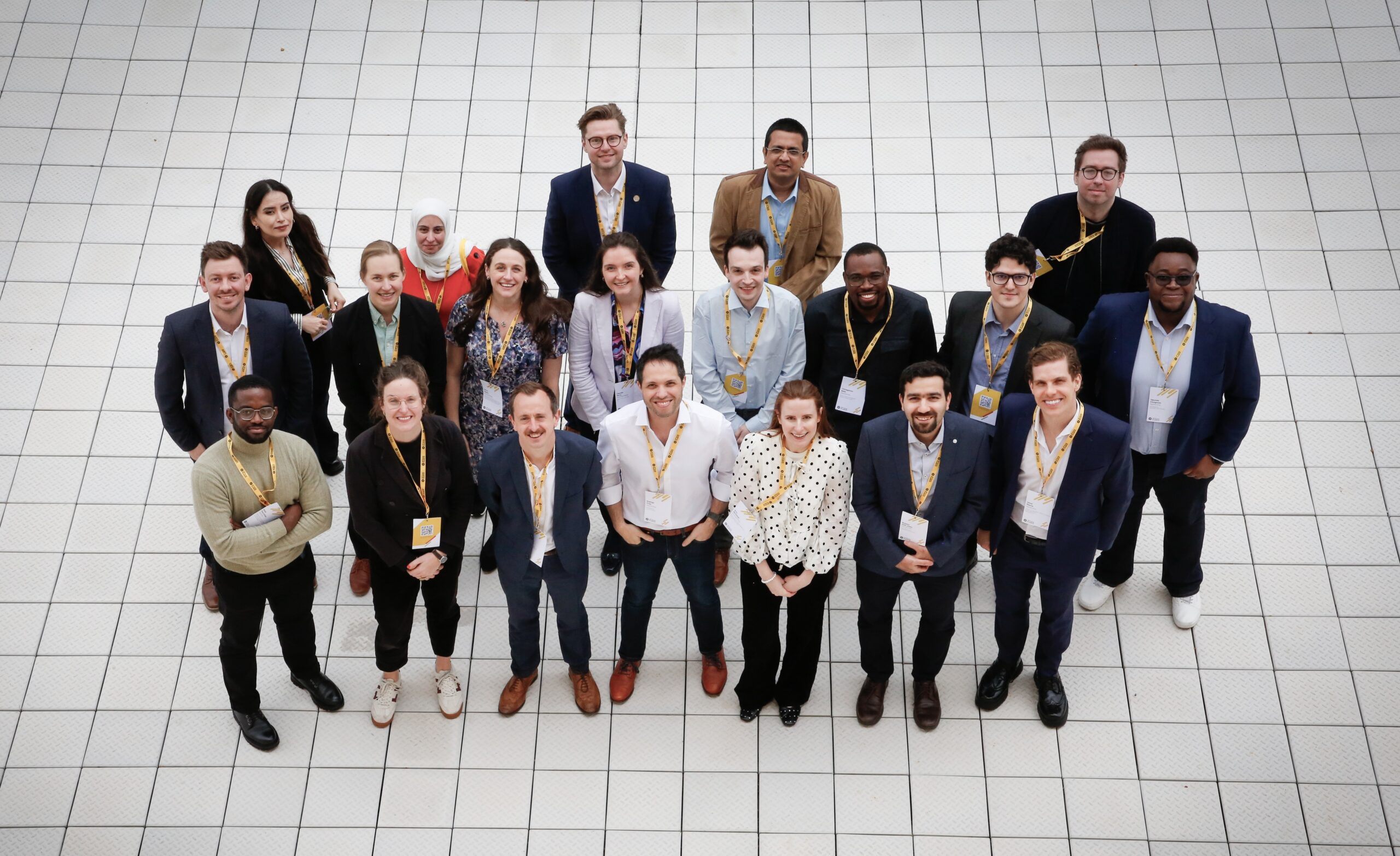 Overhead group photo of around 20 new members attending the All Members' Meeting wearing UK Young Academy lanyards and standing together on a white tiled atrium floor.